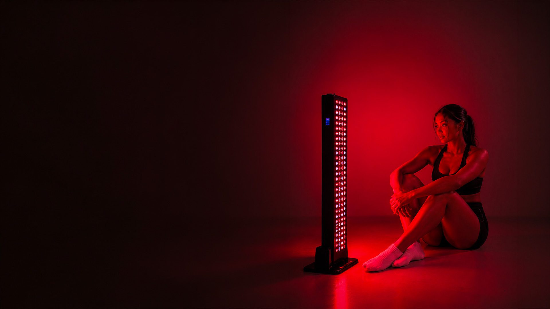 A woman seated on the floor facing a glowing Longevity Labs red light panel